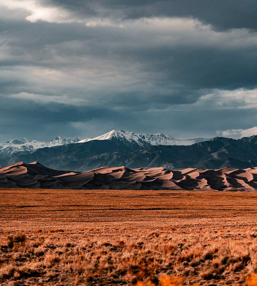 brown field near snow-covered mountains under cloudy sky.