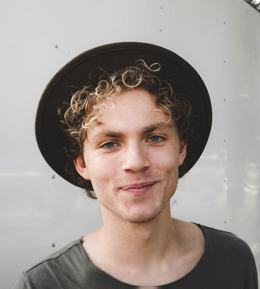 man wearing a flat-brimmed hat with short blonde curly hair and a no-teeth smile.