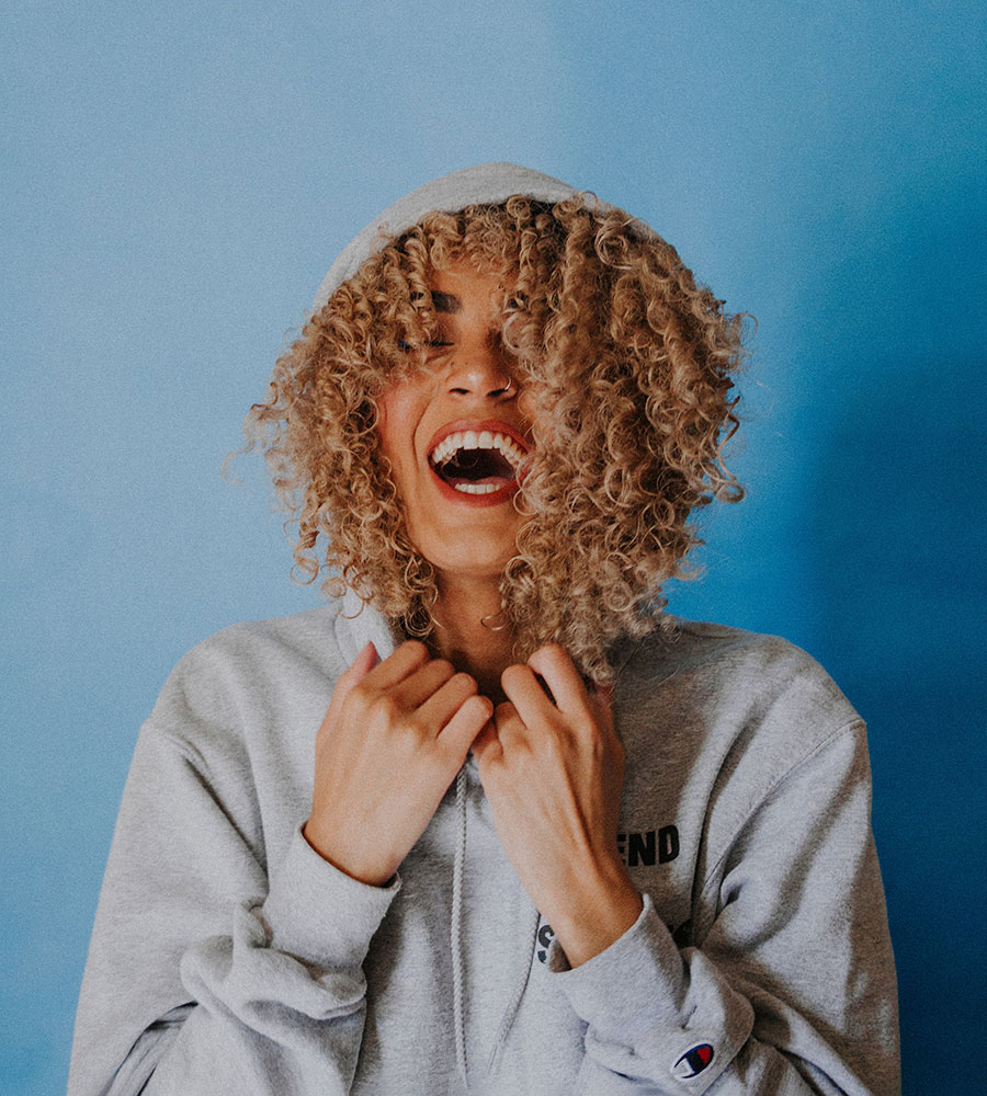 woman wearing a grey hoodie with blonde spiral curled hair laughing with her eyes closed.