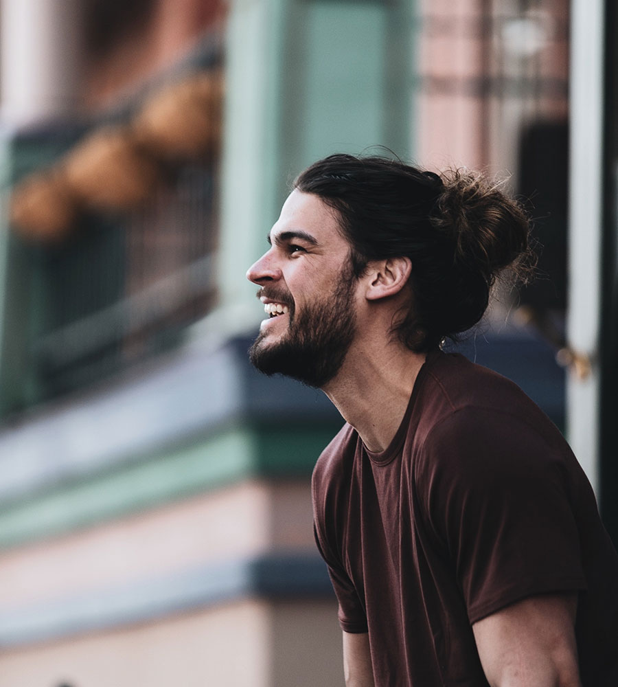 man with a beard and man-bun standing on a sidewalk looking towards the street and laughing.