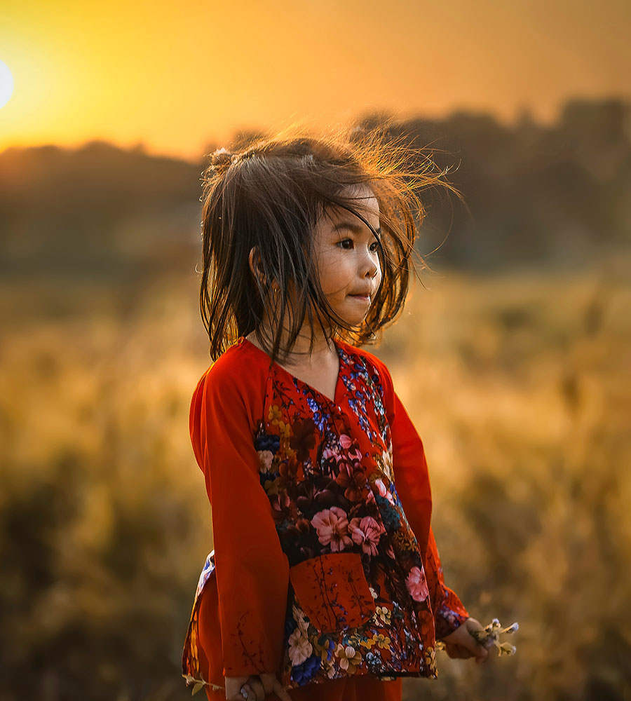 little girl in a red floral dress standing in a field of cattails with the wind blowing her hair.